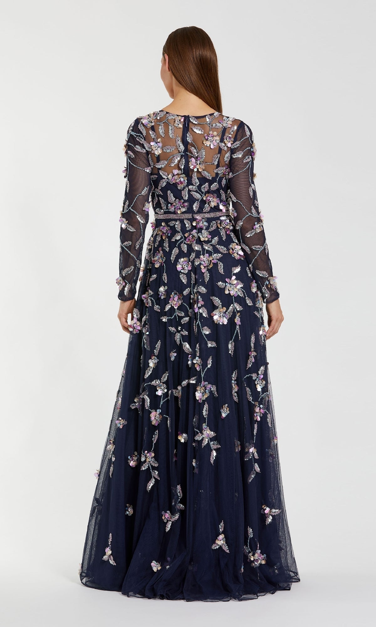 A woman stands facing away in the Mac Duggal 6183 Long Sleeve Beaded Formal Gown, a floor-length navy blue dress featuring sheer fabric and intricate floral embroidery.